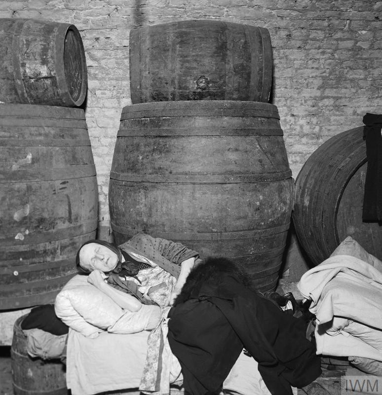 A woman sleeps on a bed made on top of a row of barrels in the cellar of a wine merchant's in East London in 1940.jpg